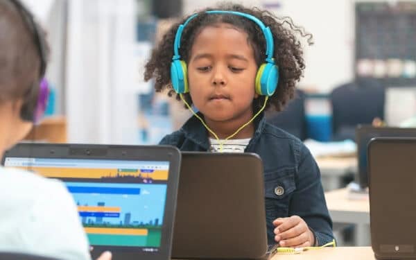 Child sitting at a laptop across from a friend with blue headphones on