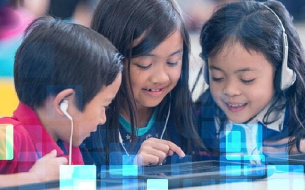 Three children learning in a group around a single tablet with headphones on